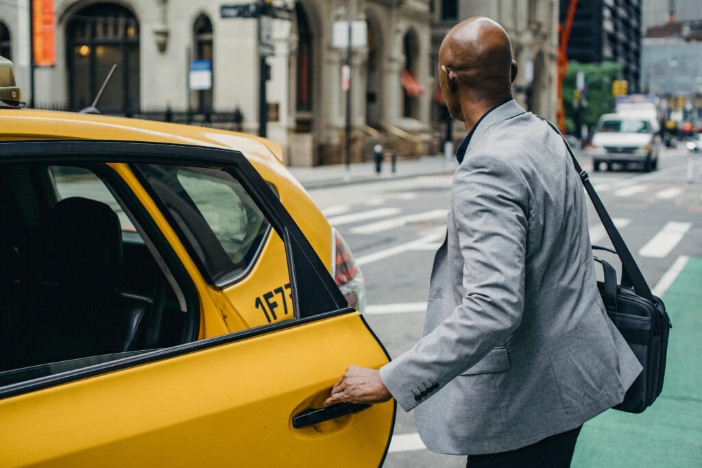 A businessman in a gray suit opens the door of a yellow taxi on a bustling city street, ready for his daily commute.